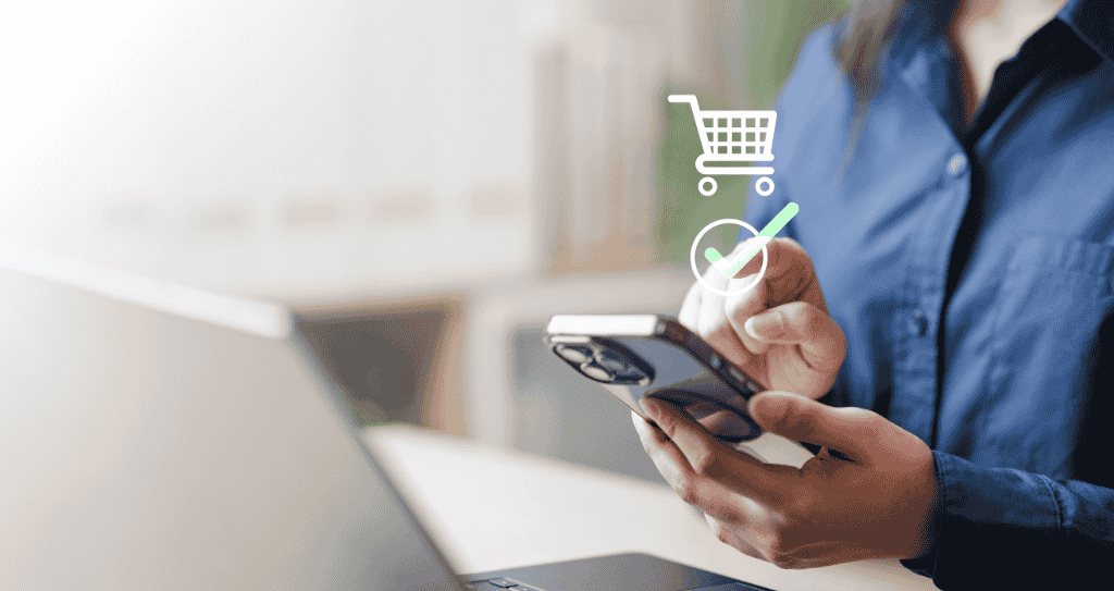 Homem utilizando celular para realizar checkout transparente de compra, em frente a um notebook, enquanto um ícone digital de carrinho de compra salta da tela do celular.