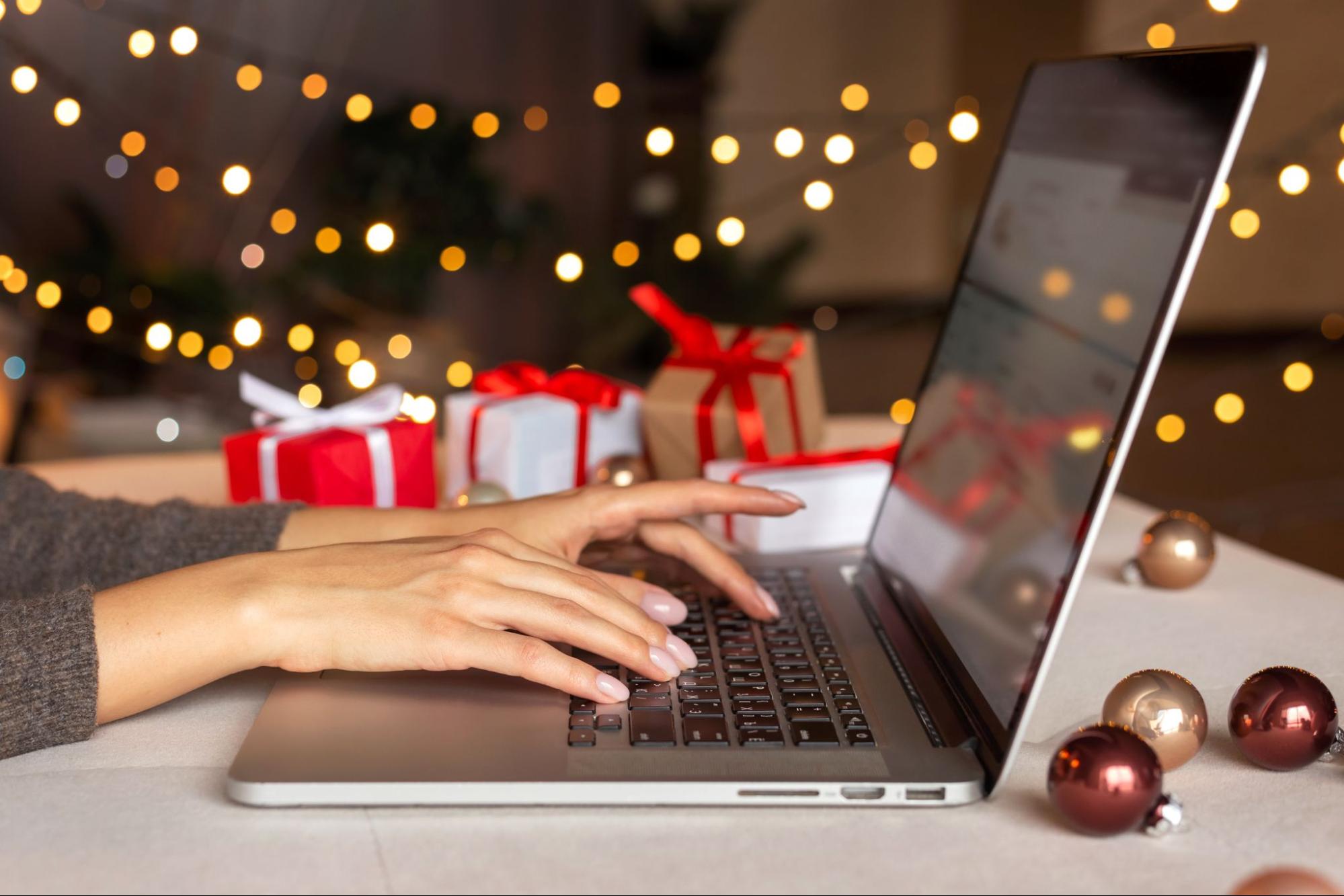 Pessoa digitando em um notebook cercado de presentes e luzes festivas, representando como criar promoções de natal em lojas virtuais e estratégias de vendas online.
