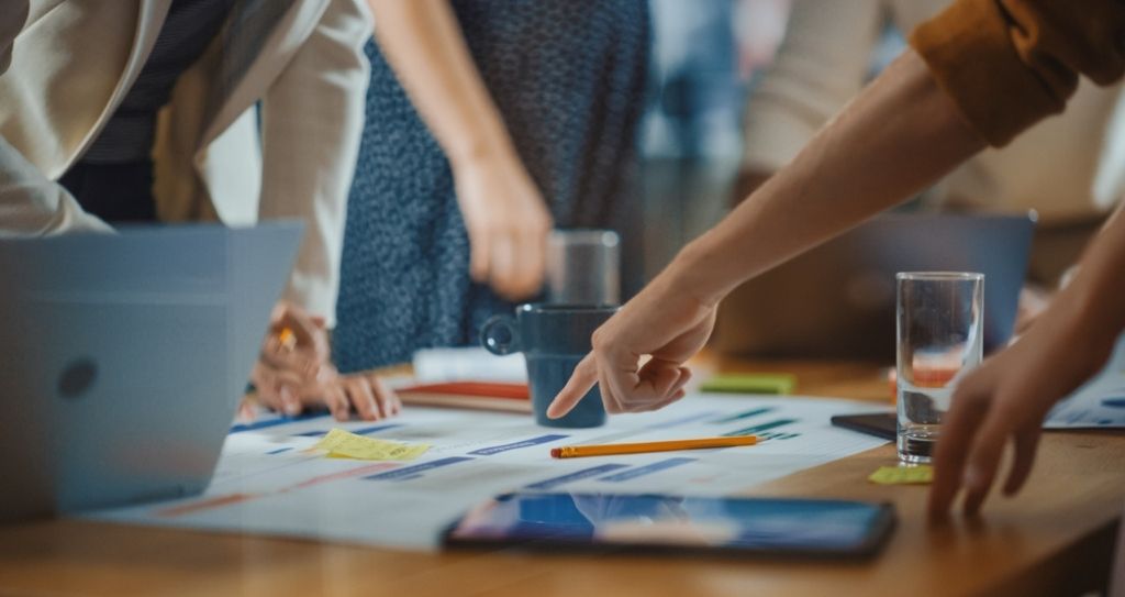Detalhe de mãos de diversas pessoas trabalhando em conjunto e apontando para cartazes e papéis com dados e outros detalhes importantes para gestão de crise.