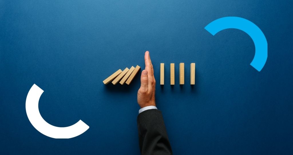 Detalhe de mão e antebraço, vestindo terno, bloqueando peças de dominó em sequência de serem atingidas por outras pedras próximas que estão caindo.