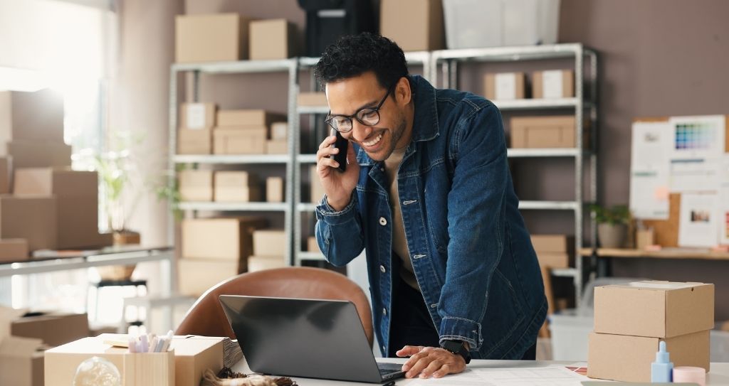 Um homem sorridente usando um celular enquanto trabalha em seu computador em um pequeno escritório, rodeado por caixas. A imagem ilustra a importância das melhores ferramentas para um pequeno negócio ao conectar comunicação e produtividade no dia a dia.