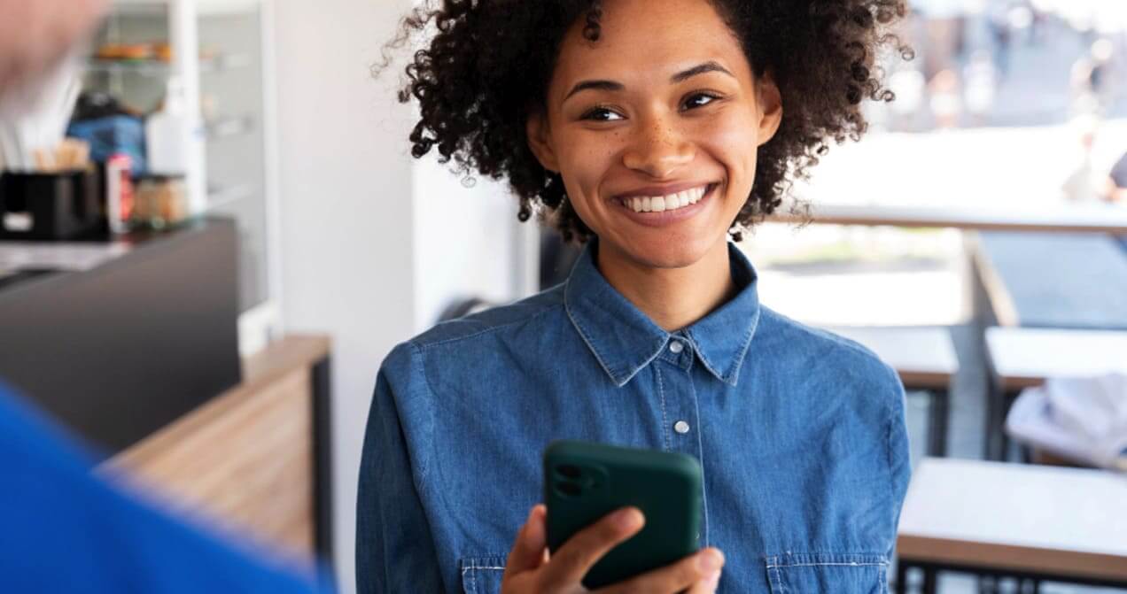 Empreendedora sorrindo ao aproximar smartphone em maquininha de cartão para pagamento.