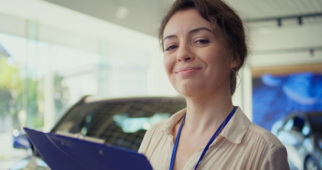 Mulher sorrindo com prancheta e crachá azul em concessionária, representa gerente de contas Cielo.