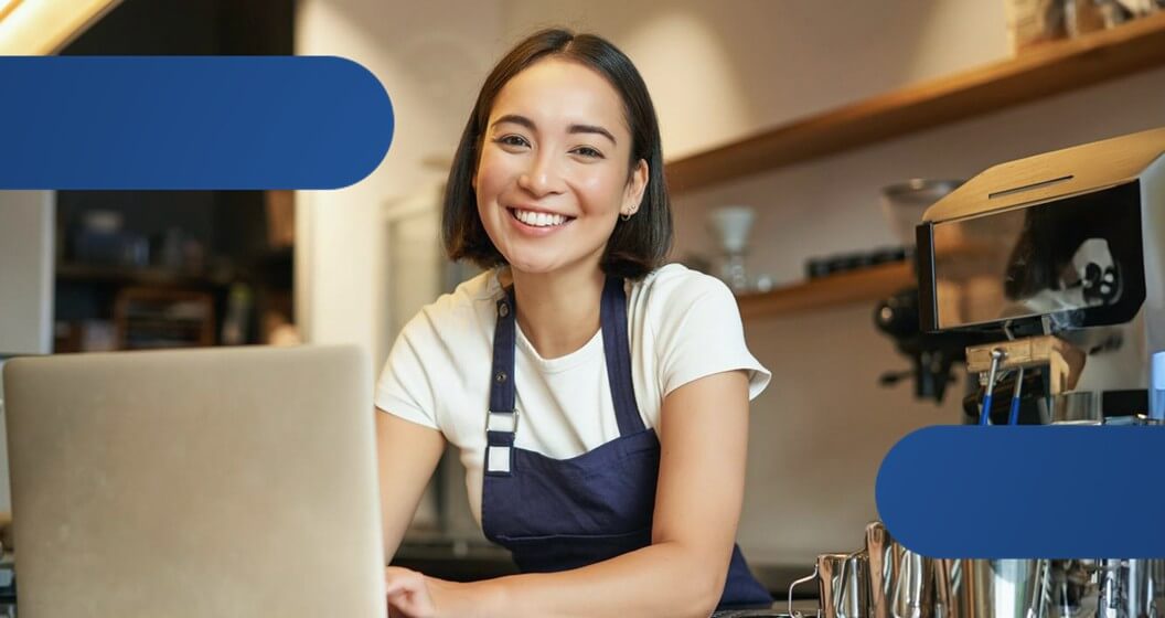 Mulher sorrindo para a câmera com notebook, onde pesquisa boas taxa de maquininha.