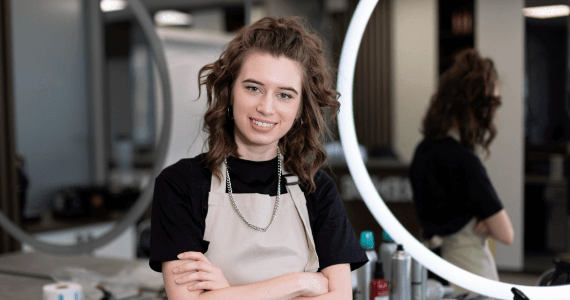 profissional de salão de beleza com avental sorrindo de braços cruzados em frente ao espelho.
