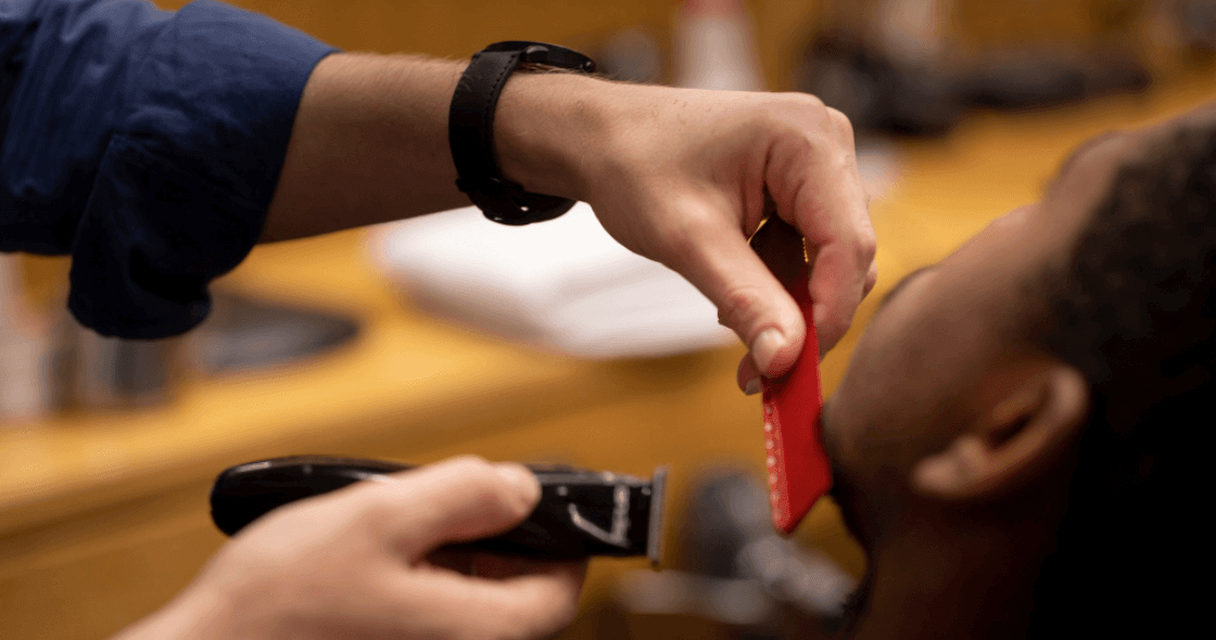 barbeiro usando máquina de corte e pente vermelho para aparar a barba de um cliente.