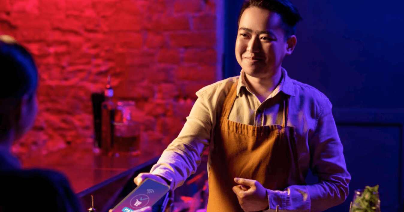 bartender sorridente oferece maquininha para pagamento por aproximação no balcão de um bar.