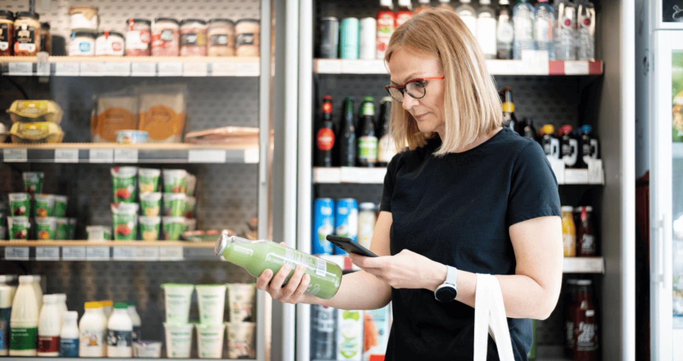 Cliente em supermercado mexendo no celular com uma mão e segurando uma garrafa de vidro com a outra.