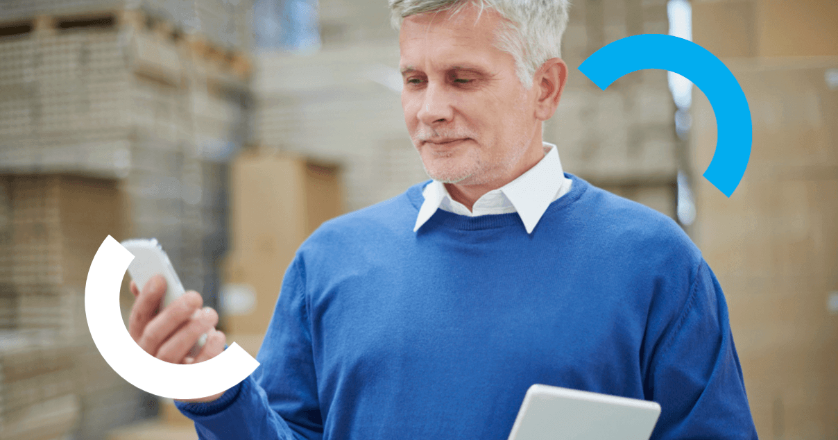 empreendedor de meia-idade com suéter azul consultando smartphone e tablet em galpão de estoque.