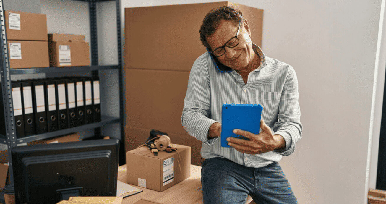 empreendedor de meia idade fala ao telefone e usa tablet vermelho em depósito com caixas e pedidos.