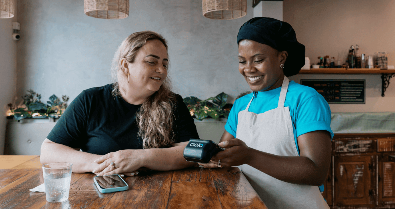 empreendedora em seu restaurante recebendo pagamento de cliente com maquininha Cielo.