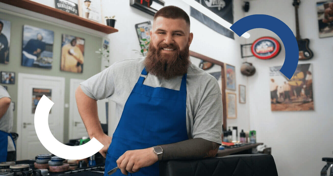 barbeiro sorridente de avental azul posando em barbearia consultando termos de pagamento para seu negócio.