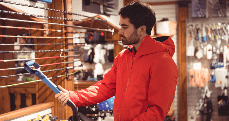 Homem de jaqueta vermelha observa uma pá em uma loja de materiais para construção.