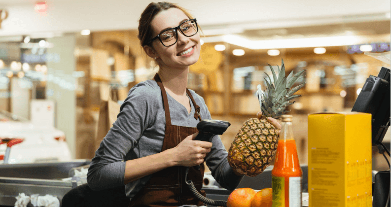 Mulher com óculos, trabalhando como caixa de supermercado, segurando um abacaxi e um leitor de código de barras.