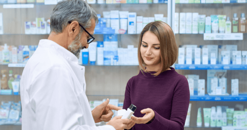 Paciente recebendo orientação de um farmacêutico em uma farmácia, com produtos farmacêuticos ao fundo.