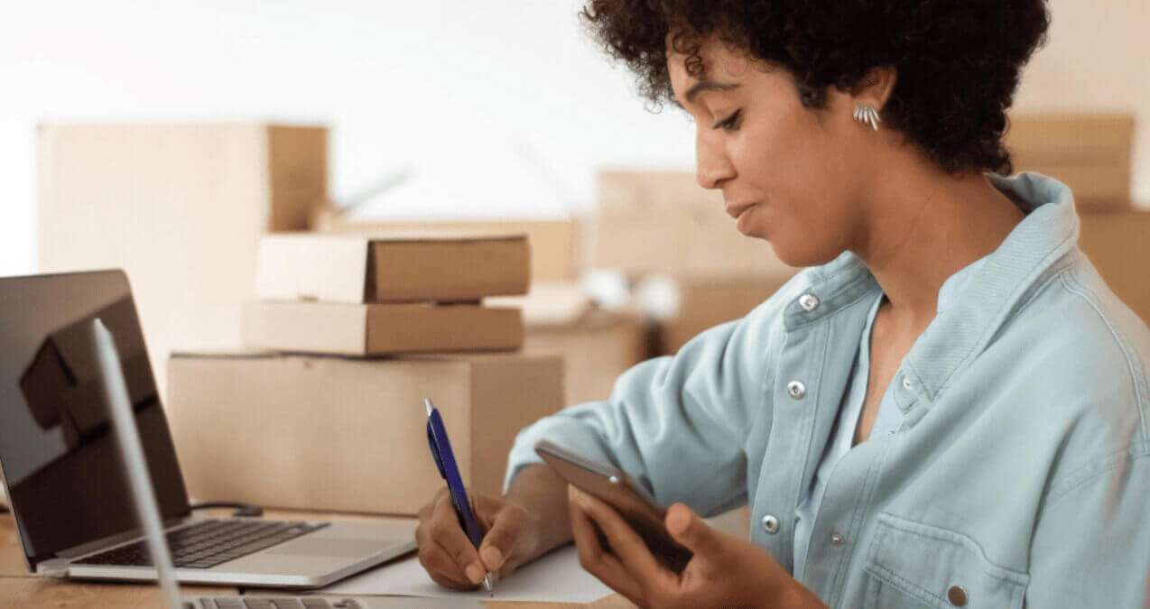 empreendedora consulta glossário de pagamentos no celular enquanto anota informações com notebook e caixas ao fundo.