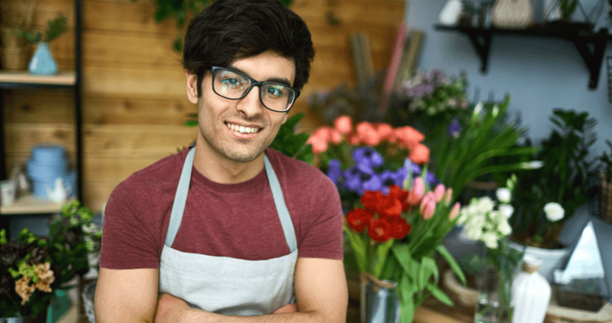 florista jovem de avental sorrindo com braços cruzados em loja de flores coloridas.