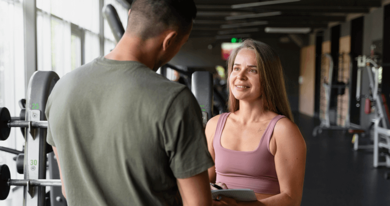 Personal trainer conversando com cliente em uma academia com aparelhos ao fundo.