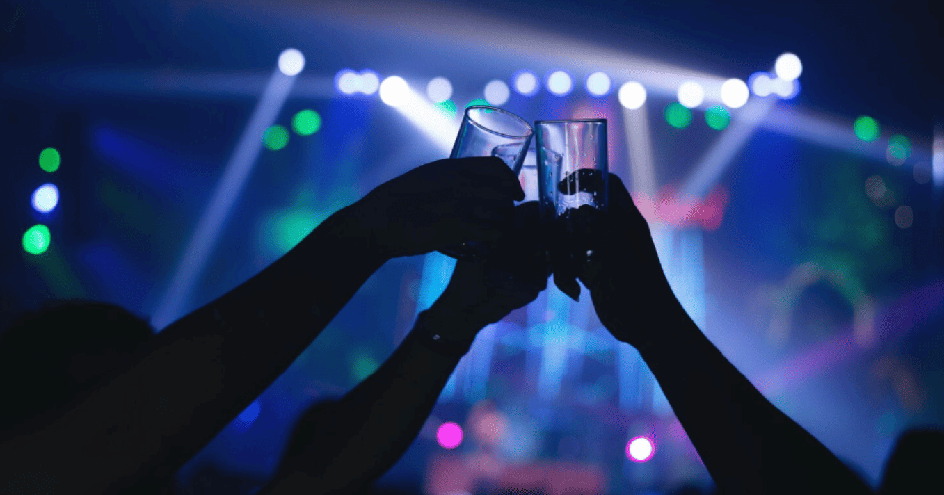 duas mãos erguidas fazem um brinde com shots em meio às luzes coloridas de uma balada.