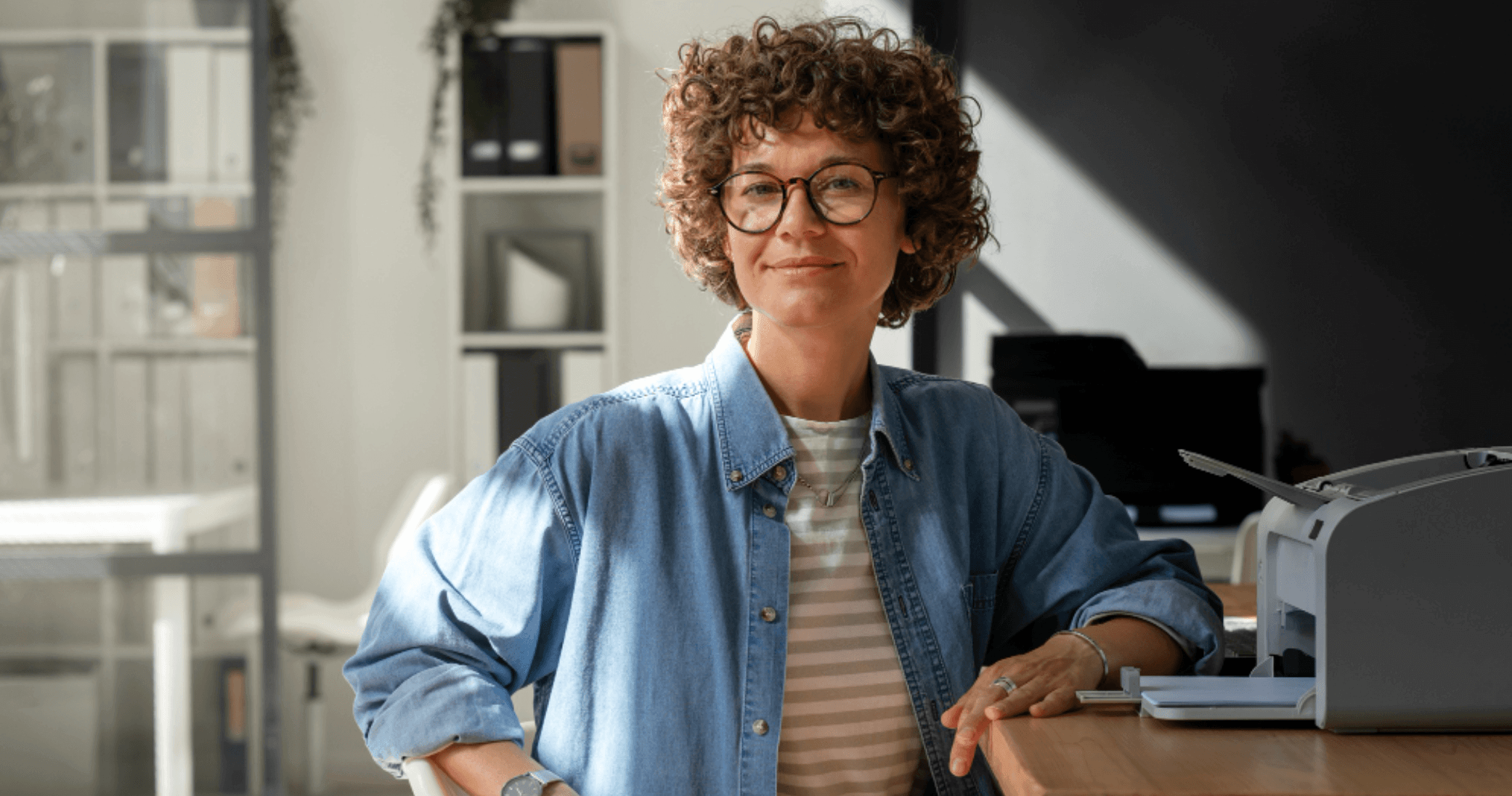 advogada ou contadora sorrindo apoiada na mesa em escritório com impressora ao fundo.