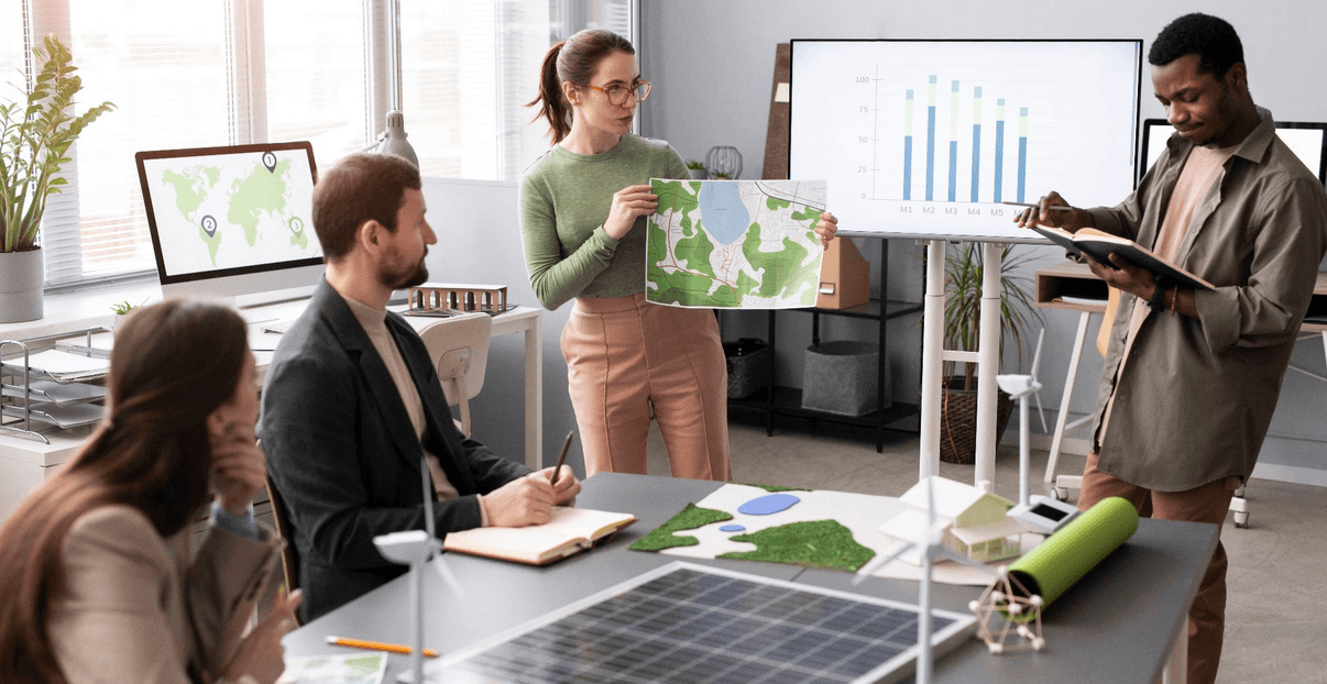 Equipe em reunião no escritório, com uma mulher apresentando um mapa em papel e outros colegas analisando gráficos em monitores, cenário de planejamento e estratégia.