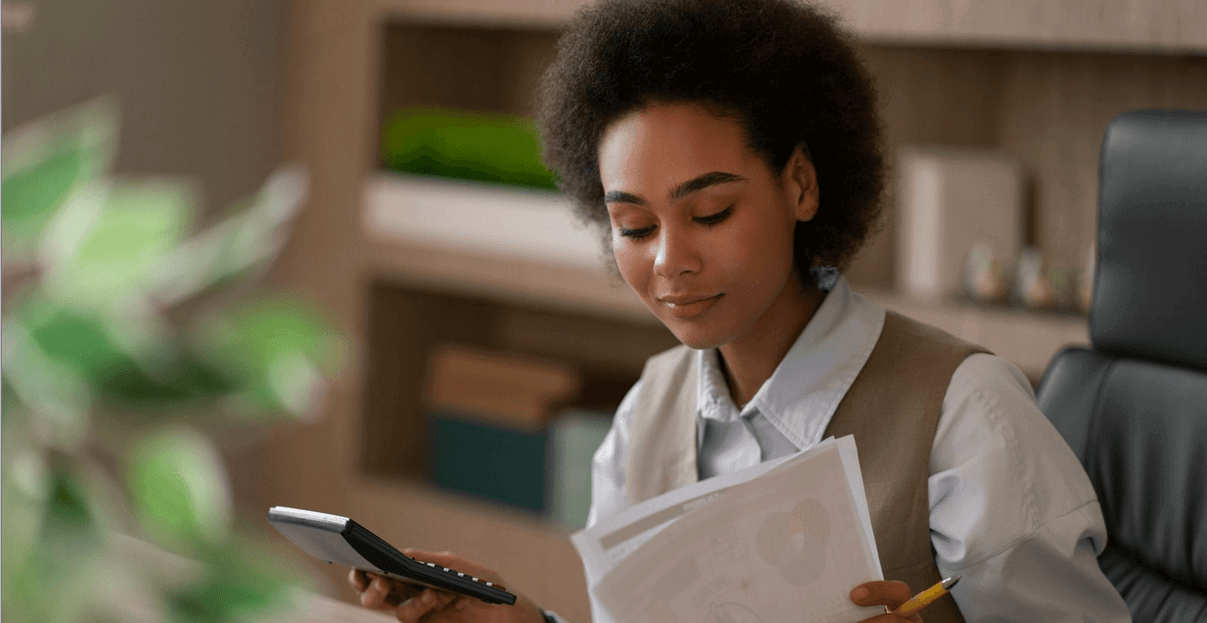 Jovem profissional sentada em ambiente de escritório, com cabelo afro, lendo documentos e usando um celular, com caixas e pastas ao fundo.