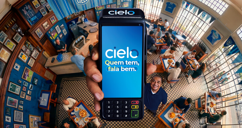 Maquininha Cielo com mensagem “Quem tem, fala bem” em destaque na tela, sendo erguida pelo ex-jogador Vampeta. Ele está em restaurante movimentado no Brasil, mostrando clientes em mesas e soluções de pagamento no dia a dia de pequenos negócios. A imagem faz parte da nova campanha da Cielo.