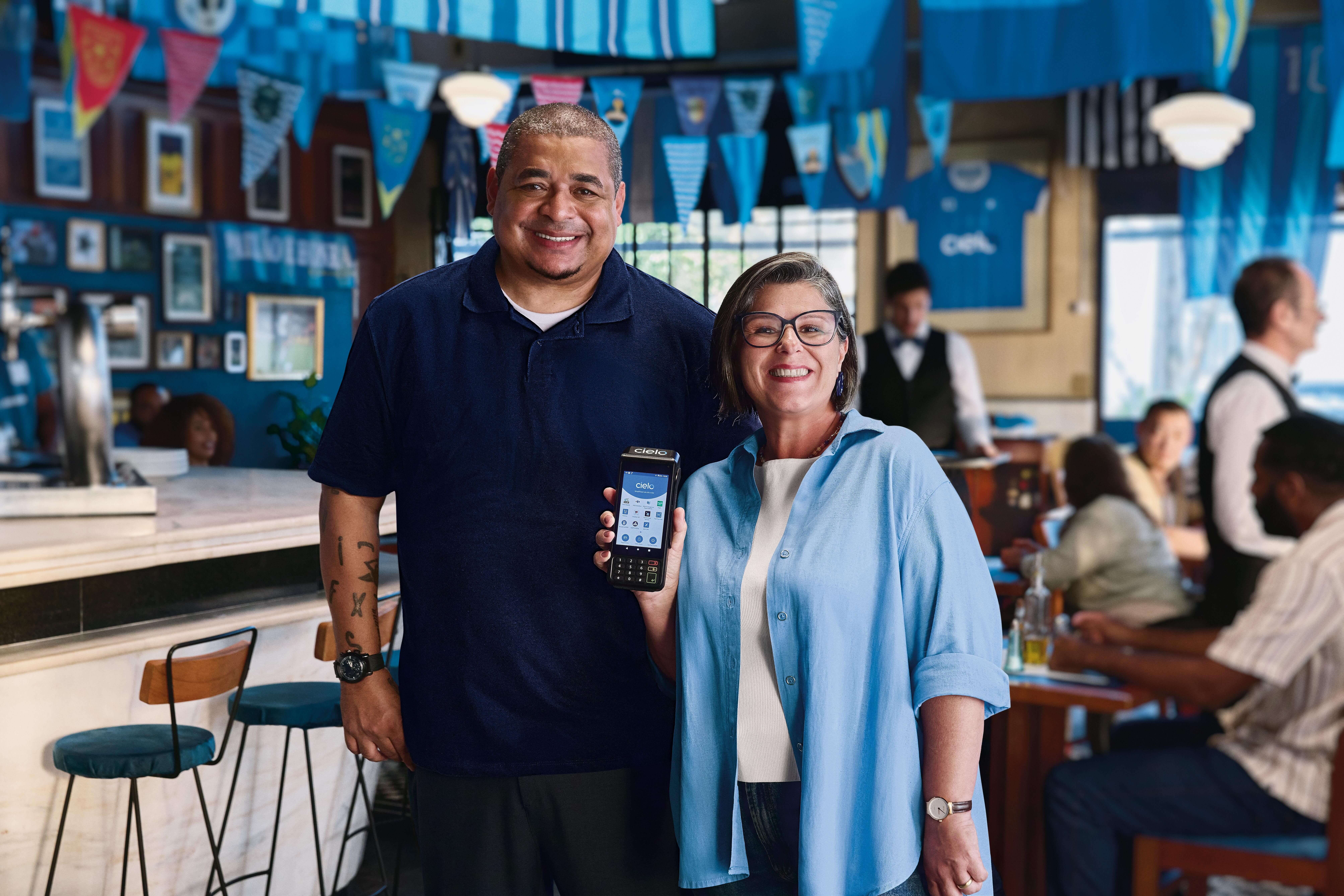 O ex-jogador Vampeta, ao lado de Cíntia Menegazzo, dona do Restaurante Tia Lourdes e cliente Cielo, posando no ambiente do estabelecimento comercial de Cíntia. Pode-se ver clientes ao fundo, destacando soluções de pagamento digital, gestão do negócio e vendas no dia a dia.
