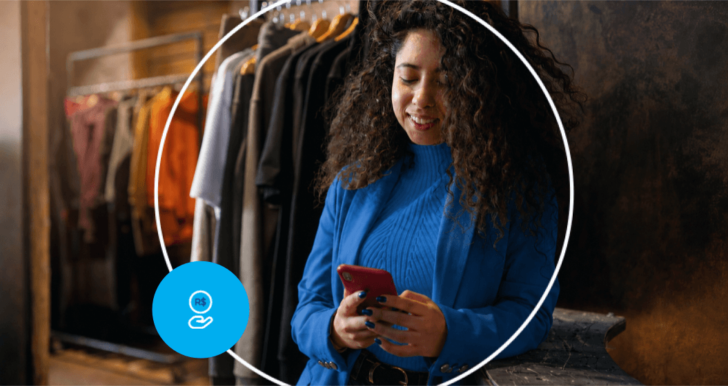 Mulher olhando para o celular em frente a um cabideiro com várias roupas.
