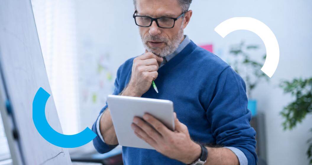 Homem de óculos e suéter azul, olha pensativo para um tablet que segura em frente a um quadro branco.