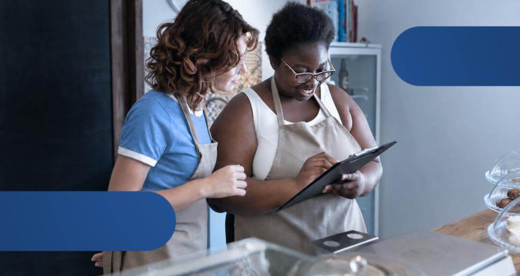 Duas colaboradoras usando um tablet para consultar informações durante o atendimento em um balcão comercial.