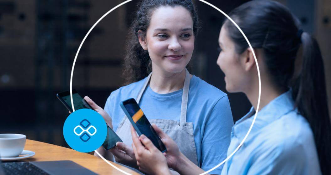 Duas atendentes conversando enquanto utilizam smartphones para demonstrar um sistema de pagamento digital em um balcão de atendimento.