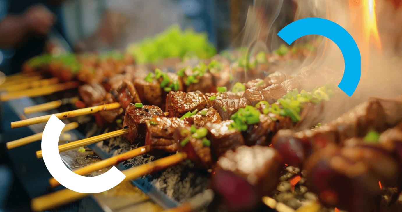 Espetinhos de carne assando na grelha com fumaça e chamas ao fundo, representando preparo de churrasco e comida de rua.