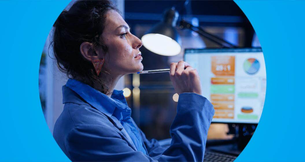 Mulher analisa informações em um monitor de computador enquanto segura uma caneta próxima ao rosto, em ambiente de trabalho.