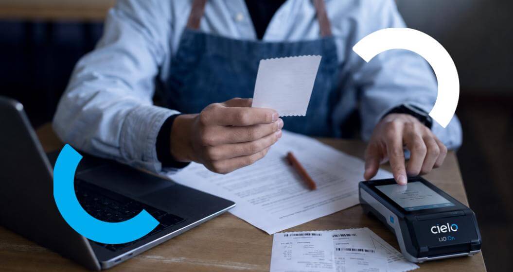 Homem de avental segura um pequeno recibo e toca na tela de uma máquina de cartões Cielo LIO On sobre uma mesa de madeira.Ao seu redor estão um notebook aberto, papéis e caneta, com grafismos circulares em azul e branco decorando as laterais da imagem.