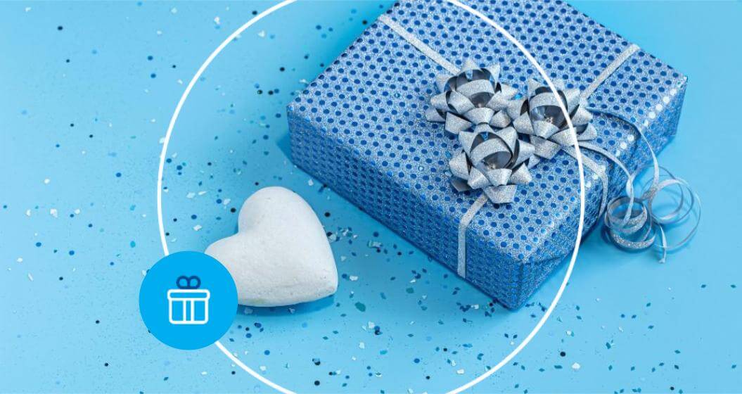 Caixa de presente azul brilhante com laços prateados ao lado de um coração branco sobre fundo azul com glitter. A imagem é emoldurada por um círculo branco e traz um ícone redondo azul com o desenho de um presente no canto esquerdo.