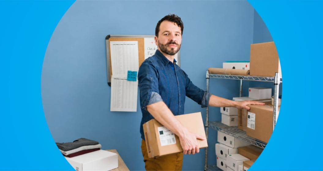Um homem de barba e camisa jeans segura uma caixa de papelão ao lado de uma estante de metal cheia de encomendas, em uma sala com parede azul.A cena é apresentada dentro de uma moldura circular centralizada sobre um fundo azul-claro brilhante.