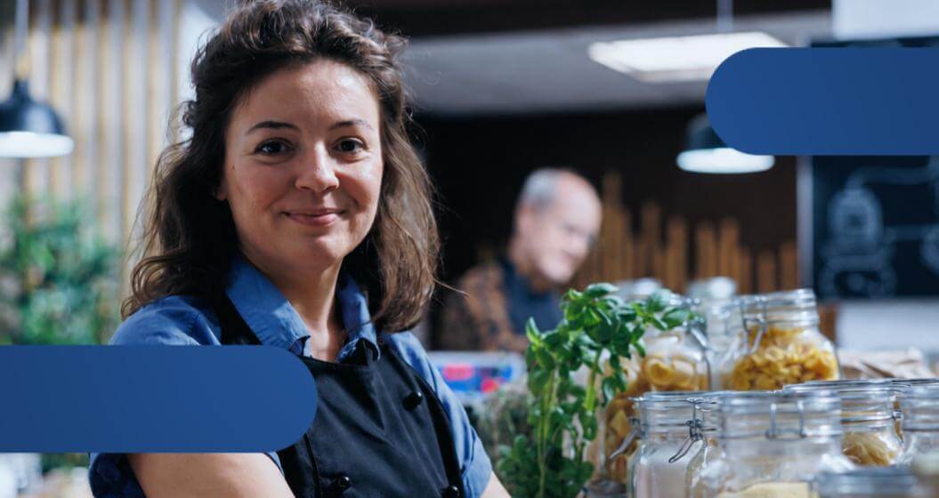 Uma mulher sorridente de avental preto e camisa azul posa em um estabelecimento comercial com potes de vidro cheios de alimentos em primeiro plano.Ao fundo, um homem aparece desfocado, e a imagem possui detalhes gráficos em formato de formas arredondadas azuis nos cantos.