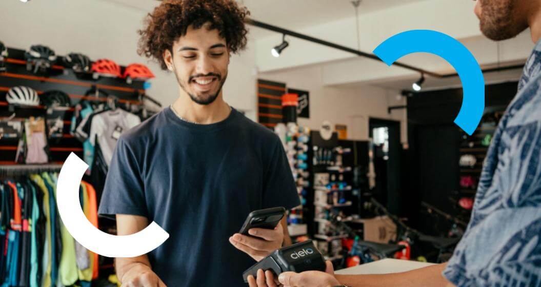 Um jovem de cabelo cacheado sorri ao aproximar o celular de uma maquininha de cartão Cielo para fazer um pagamento em uma loja de bicicletas.