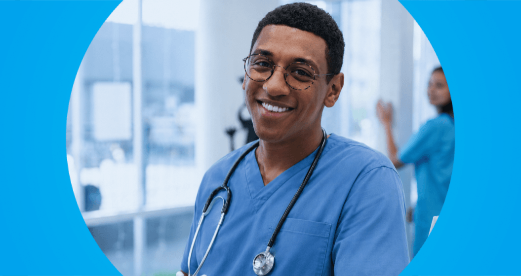Aluno de medicina sorridente, de óculos e uniforme azul, com estetoscópio no pescoço, em um ambiente hospitalar.