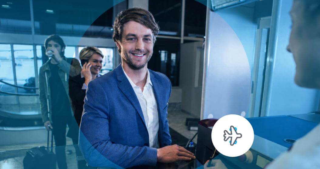 Homem sorridente, vestindo terno azul, realizando atendimento em um balcão de aeroporto, com outros viajantes ao fundo.