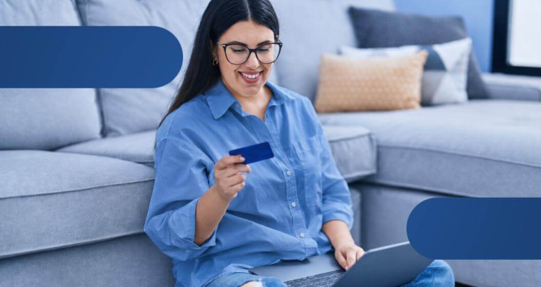 Mulher sorrindo sentada no chão usando um notebook e segurando um cartão de crédito enquanto faz uma compra online em casa.