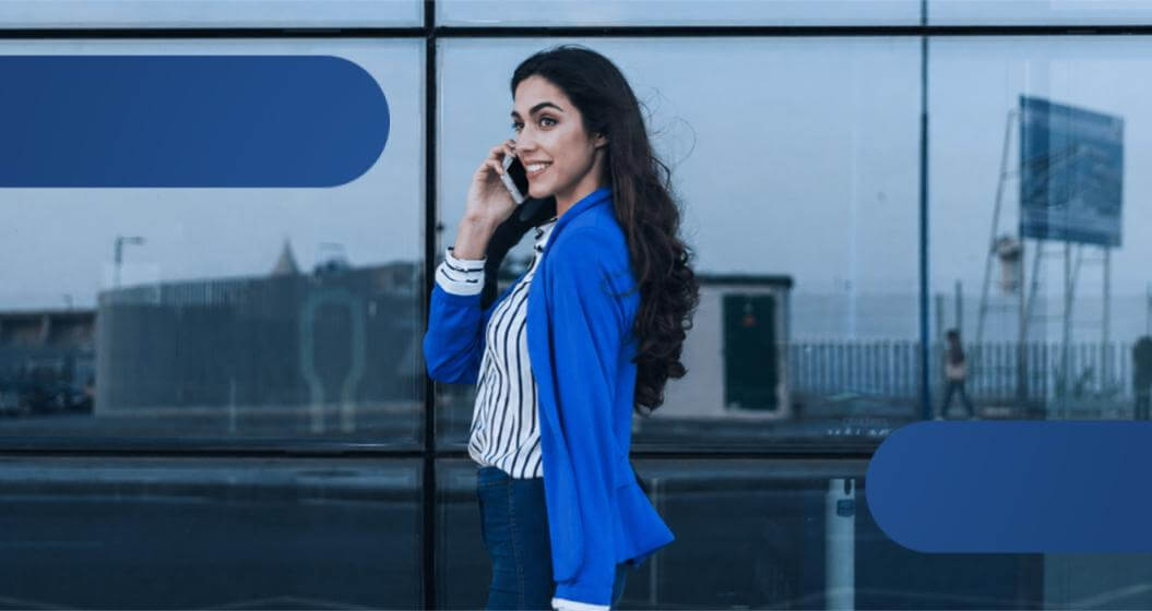 Mulher sorridente, vestindo blazer azul, falando ao celular em frente a um edifício comercial envidraçado.
