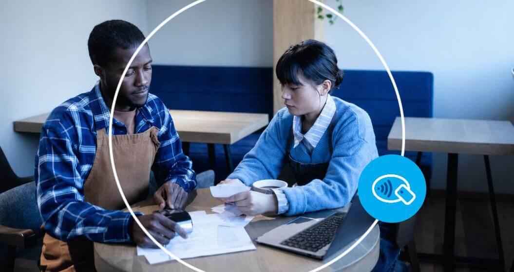 Duas pessoas analisam documentos e uma calculadora em uma mesa de trabalho, com um notebook aberto ao lado e uma xícara de café. A cena representa o planejamento financeiro de um negócio, com destaque para um ícone de pagamento por aproximação no canto inferior direito.