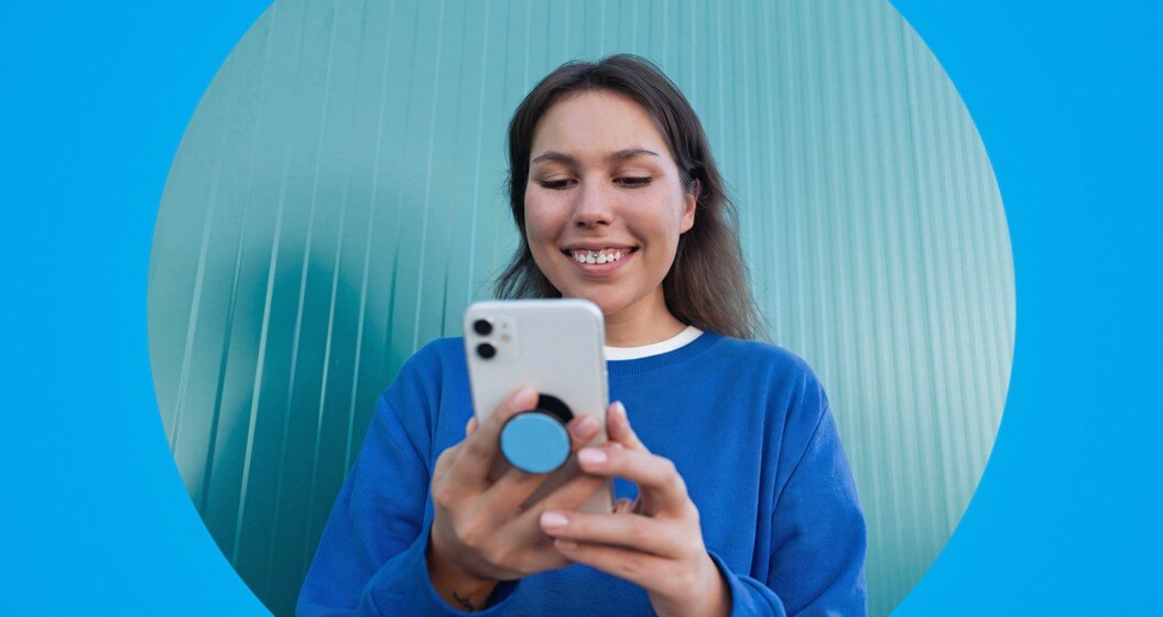 Uma mulher sorridente em moletom azul olha para um celular com um suporte de coronavírus estilizado. A imagem está contida em um círculo centralizado em um fundo azul vibrante, diante de uma parede verde-água.