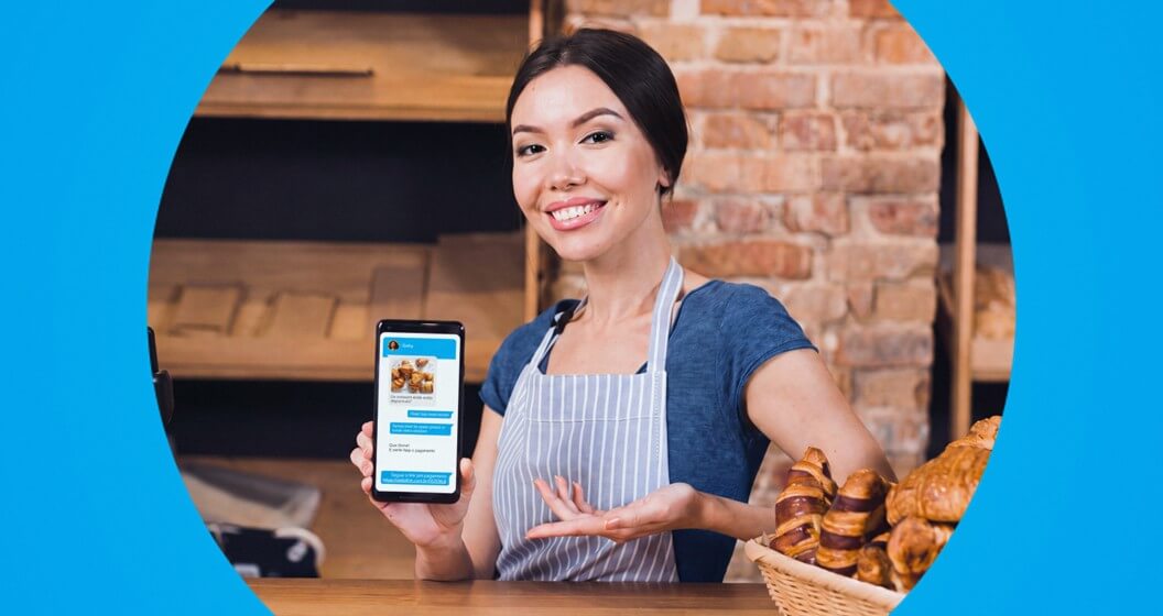 Mulher sorridente de avental listrado segura um celular exibindo uma conversa de aplicativo e gesticula para uma cesta de croissants sobre o balcão de uma padaria. A foto possui recorte circular sobre fundo azul.