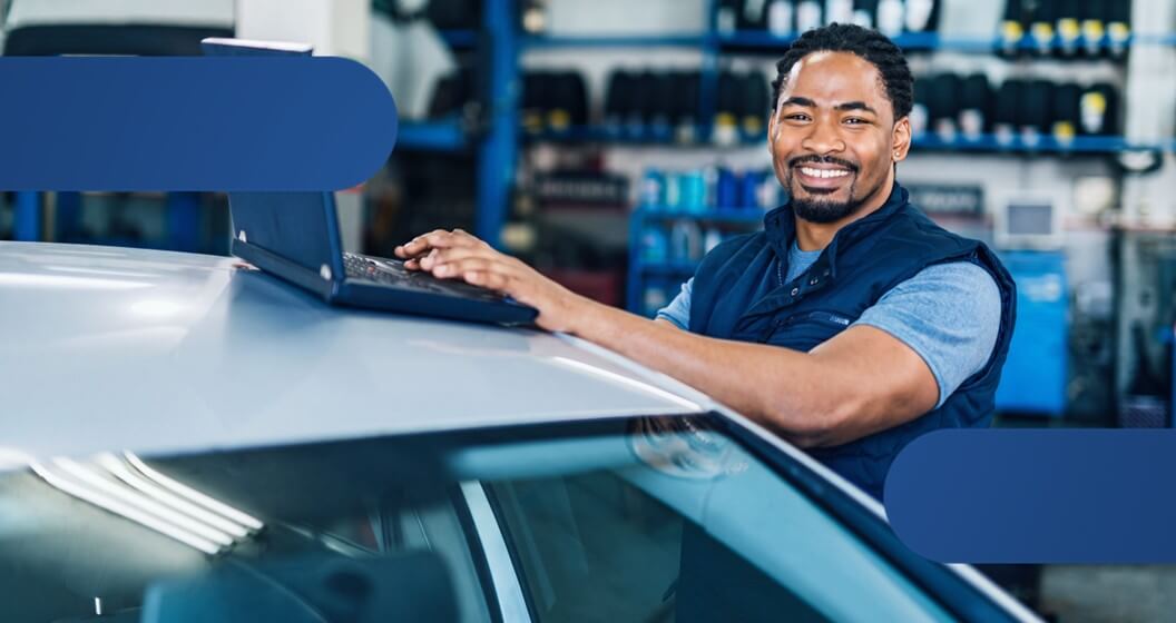 Homem negro sorridente, vestindo colete azul, apoia as mãos em um notebook aberto sobre o teto de um carro prata. Ele está em uma oficina mecânica com prateleiras ao fundo, e a imagem conta com detalhes em azul-escuro nos cantos superior esquerdo e inferior direito.