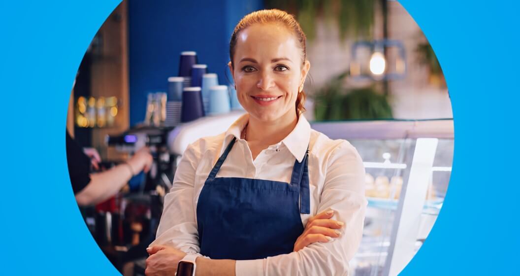 Uma mulher sorridente, vestindo avental azul e camisa branca, posa com os braços cruzados dentro de uma cafeteria. A imagem é circular e está centralizada sobre um fundo azul vibrante.