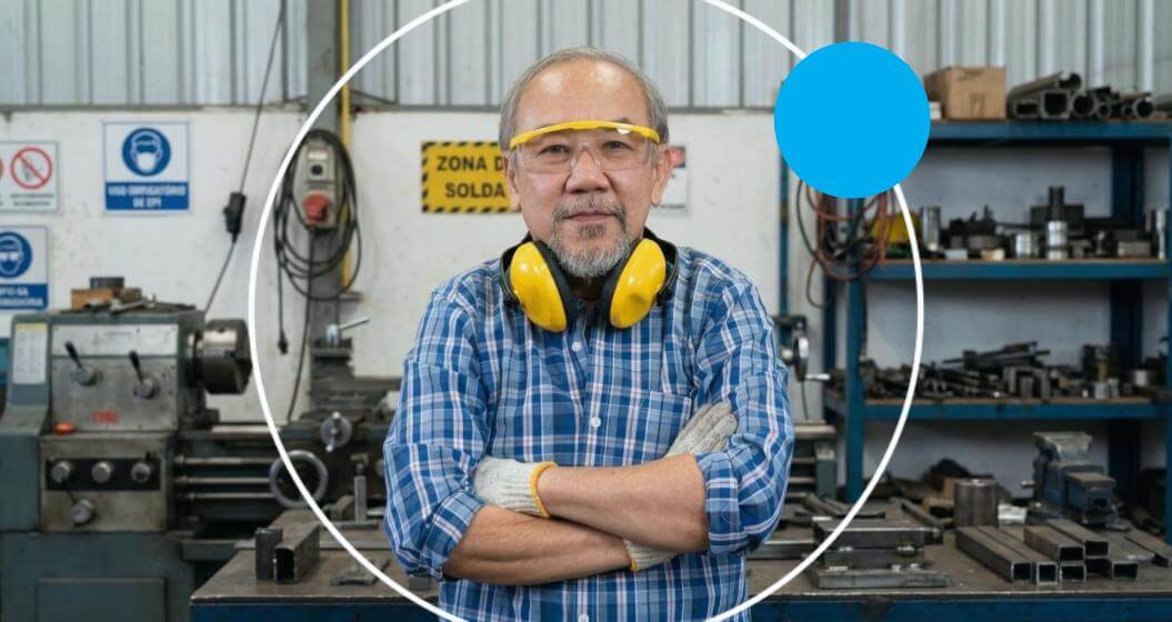 Homem asiático sênior de óculos de proteção e protetor auricular no pescoço posa de braços cruzados em uma oficina industrial. A foto tem um enquadramento circular central com borda branca e um círculo azul no canto superior direito.
