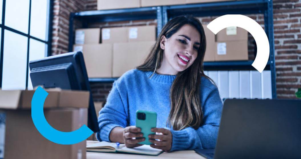 Uma mulher sorridente com casaco de lã azul segura um smartphone sobre uma mesa de escritório, com dois semicírculos coloridos sobrepostos à imagem.Ao fundo, há prateleiras com caixas de papelão e, em primeiro plano, dois laptops e um caderno aberto.